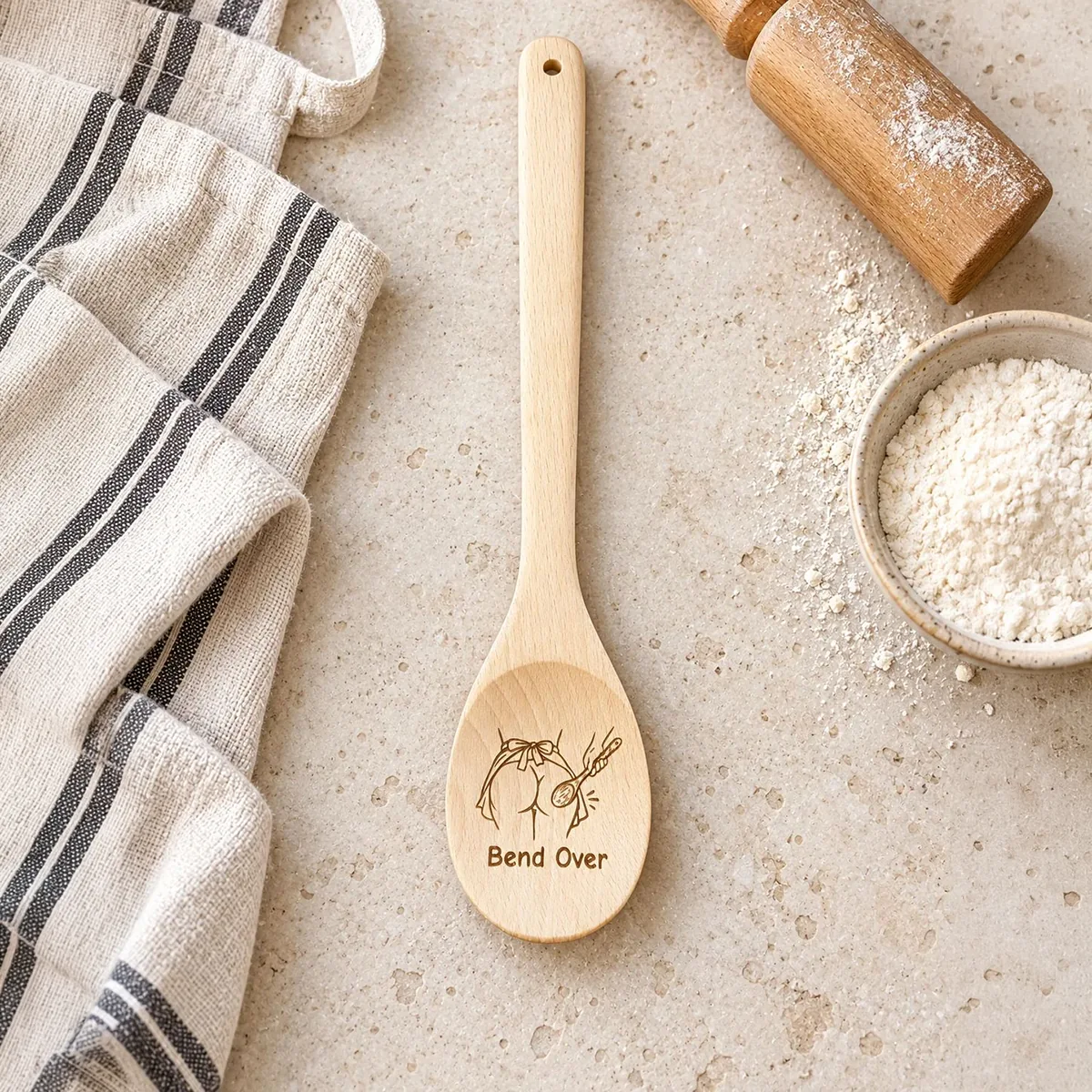 Bend Over engraved wooden spoon, on a baking tabletop with an apron and rolling pin