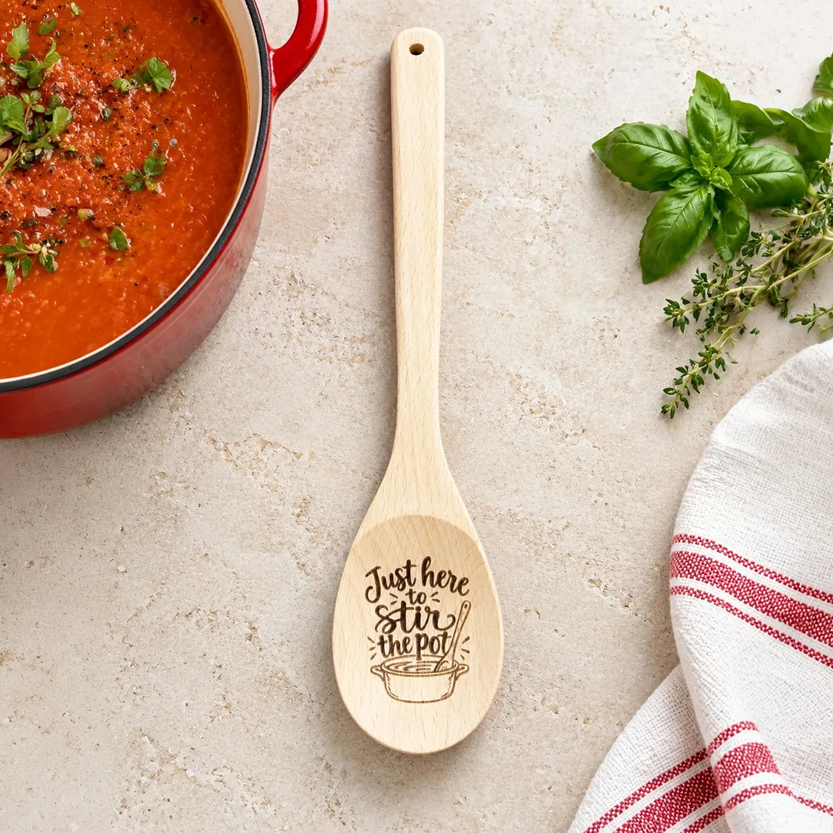 Just Here to Stir the Pot engraved wooden spoon, on a kitchen tabletop beside tomato soup