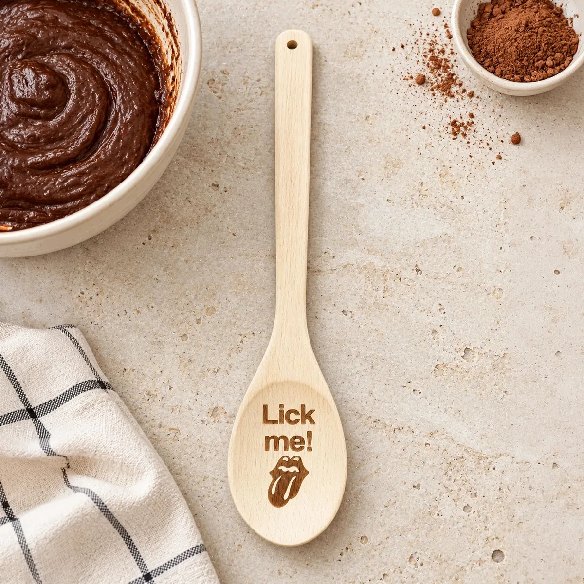 Lick Me engraved wooden spoon, on a baking tabletop beside chocolate batter