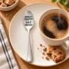 World’s Greatest Shit Stirrer Engraved Spoon beside a cup of coffee.